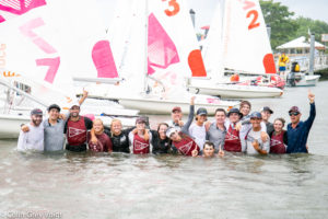 A group of smiling people in matching maroon vests stand waist-deep in water, celebrating and posing in front of sailboats. Some have raised arms; the atmosphere is joyful and festive.