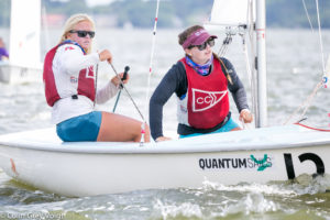 Two women in life jackets are sailing a small boat on the water. Both are focused and holding ropes, with trees and water in the background. The boat has "QUANTUM SAILS" written on it.