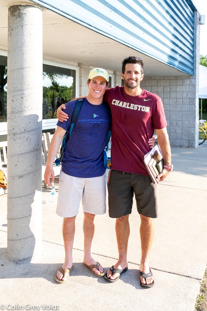 Two smiling men stand side by side outdoors, one wearing a blue shirt and khaki shorts with a cap and backpack, the other in a maroon "Charleston" shirt holding books, arm around his friend.