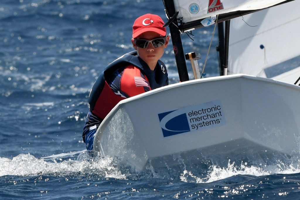 A young sailor wearing a red hat with a Turkish flag, sunglasses, and a life jacket steers a small sailboat on the water. The boat displays a U.S. flag patch and an "electronic merchant systems" logo.