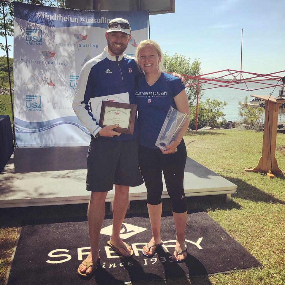 Two people stand smiling outdoors on grass, holding awards in front of a stage with a US Sailing banner. The man wears sunglasses, a hat, and shorts; the woman wears a blue shirt and leggings. A lake is visible in the background.