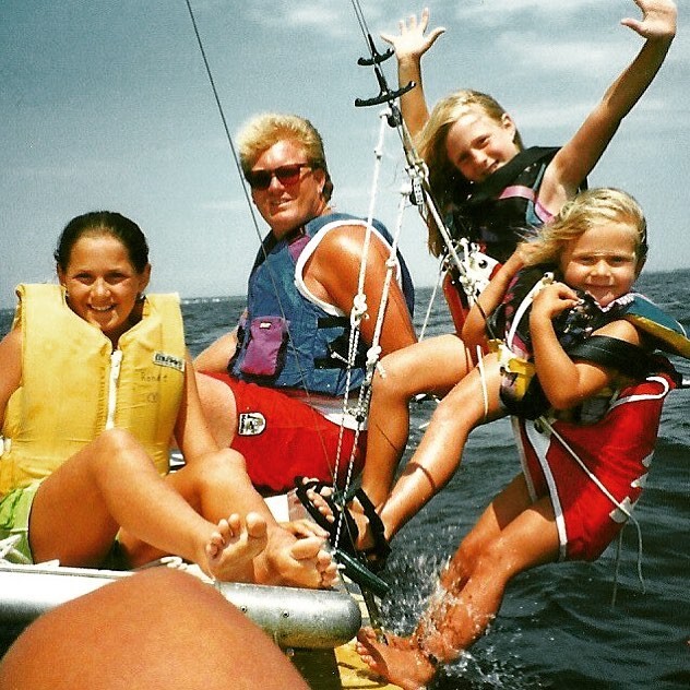 Four people, including two children and two adults, wearing life jackets, smile and pose on a small sailboat on the water. One child raises her arms joyfully, while the others relax and look at the camera. It is a sunny day.