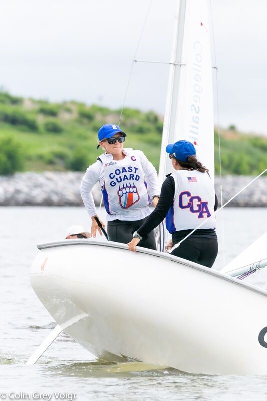 Two people wearing "COAST GUARD" vests and blue hats are standing on a white sailboat in the water, near a green shoreline, preparing to sail.