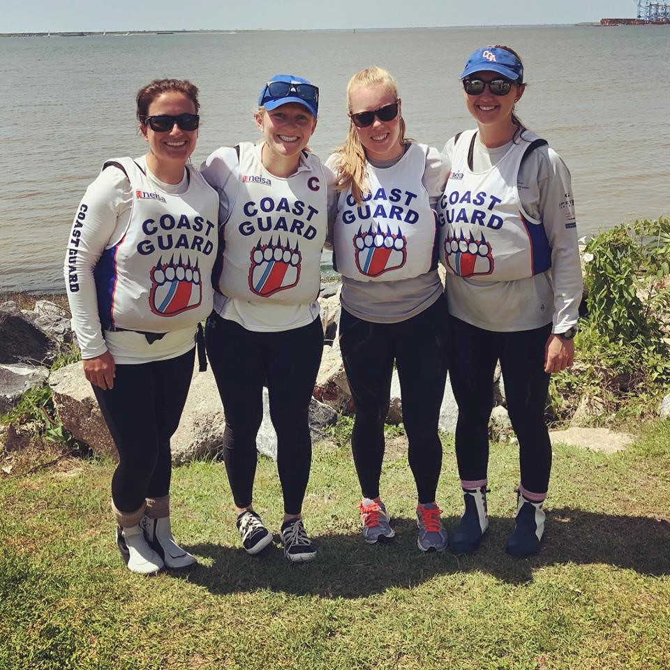 Four women wearing matching "Coast Guard" shirts and sailing gear stand together smiling by the water, with rocks and grass in the foreground and a distant shoreline in the background.