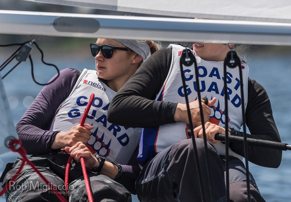 Two sailors wearing "Coast Guard" vests and sunglasses sit side by side on a sailboat, holding ropes and looking toward the left, focused on maneuvering during a sunny day on the water.