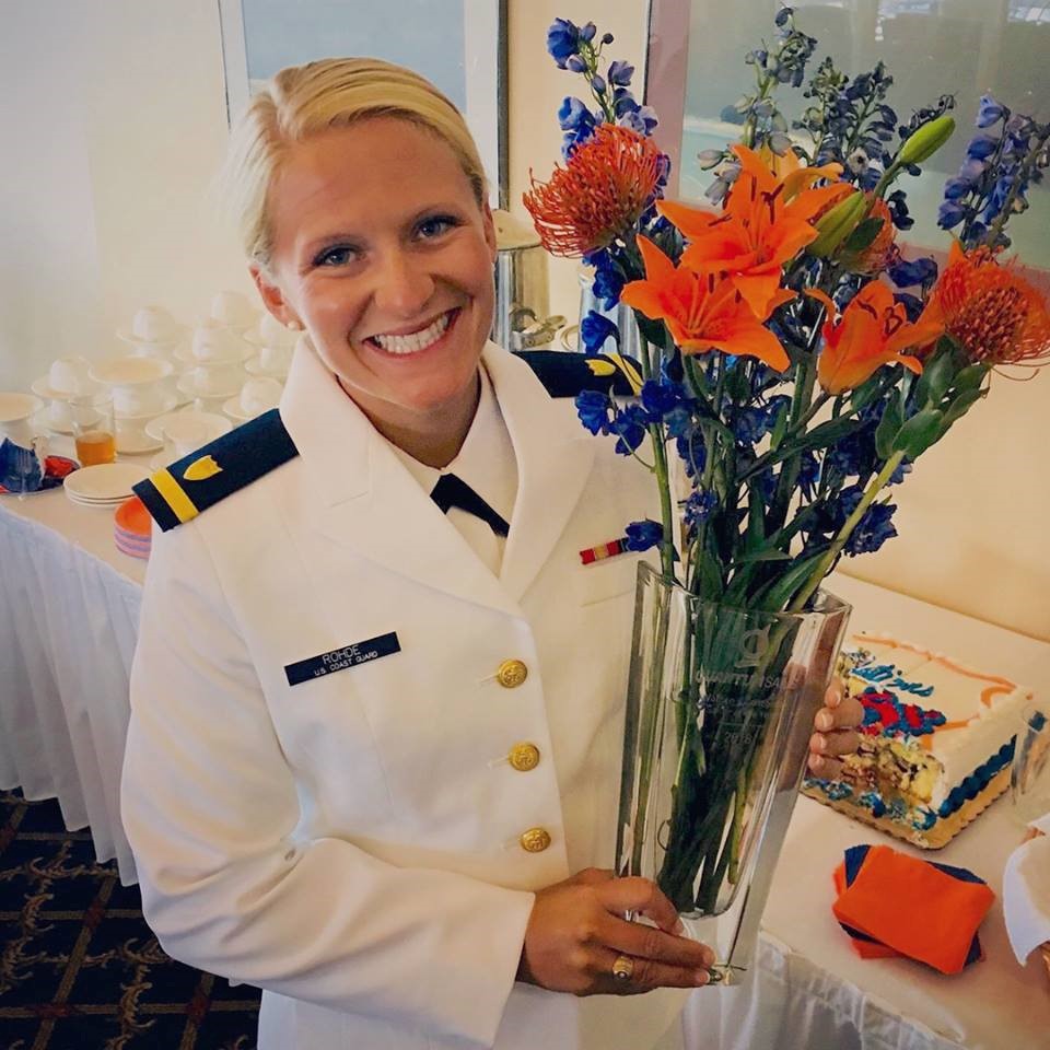 A smiling woman in a white military uniform holds a clear vase of colorful orange and blue flowers. Behind her is a table with a cake, plates, cups, and a stack of napkins.
