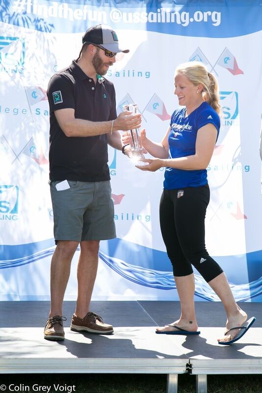 A man in sunglasses hands a glass trophy to a smiling woman in athletic clothes onstage at a sailing event, with a 'US Sailing' banner in the background.