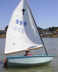 A person wearing sunglasses and a red life jacket sails a small blue and white sailboat numbered 599 on calm water, with houses and trees visible on the distant shore under a clear sky.