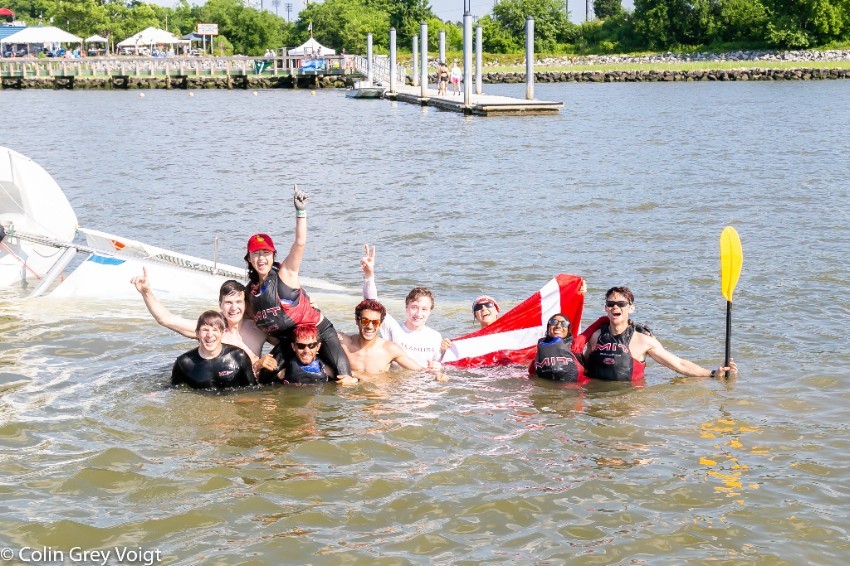 A group of people wearing swimsuits and life jackets are waist-deep in a river, smiling and raising their arms in celebration near an overturned sailboat. A dock and tents are visible in the background.