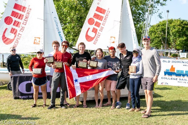 A group of nine people stand on grass in front of sailboats, holding plaques and a red and white flag with a blue star. They are smiling and posing for a photo at a sailing event on a sunny day.