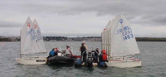 A group of people in life jackets gather on small sailboats and dinghies on a calm body of water under a cloudy sky, with a town visible in the distant background.