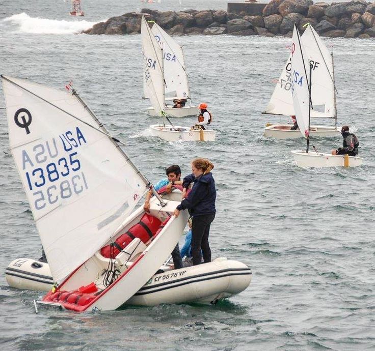 A woman helps a young sailor upright a small sailboat from a dinghy on choppy water, while other children sail similar boats nearby. Rocks and a few distant figures are visible in the background.