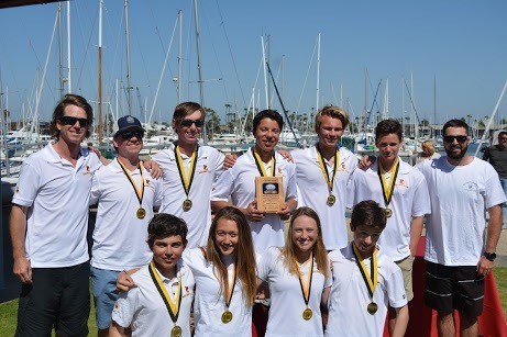 A group of ten young people wearing white shirts and medals pose together outdoors near a marina, with sailboats in the background. One person in the center is holding a plaque.