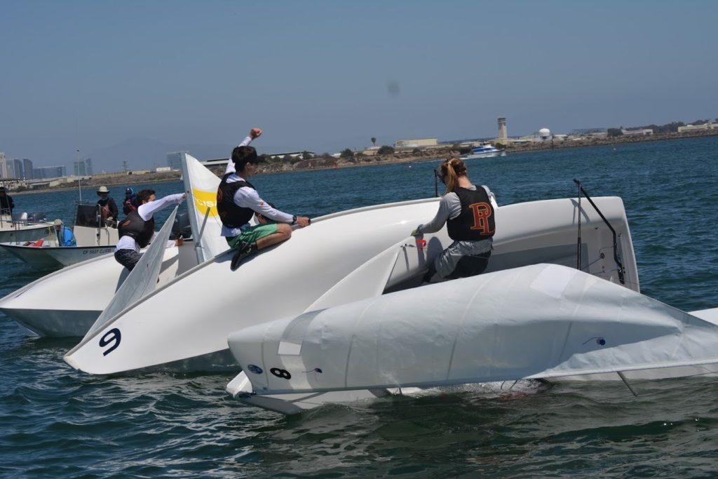 Several people in life jackets are handling two capsized small sailboats on the water, working together to right them under clear skies near a city shoreline.