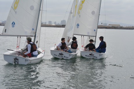Two small sailboats with four people each are sailing close together on calm water. The boats have white sails, and the city skyline is visible in the background under a cloudy sky.