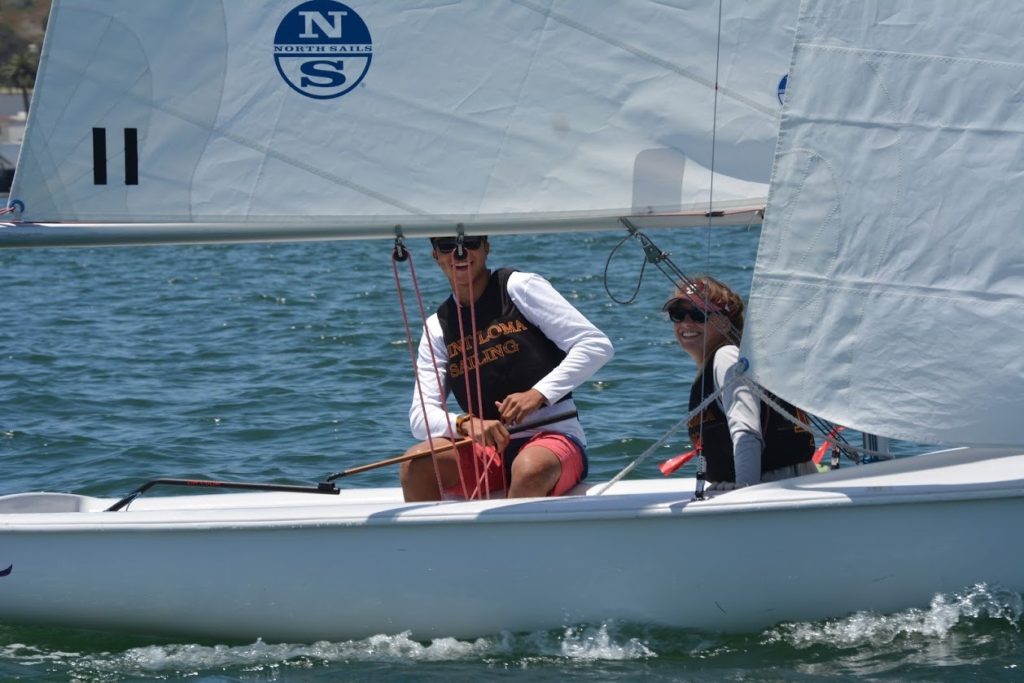 Two people wearing sunglasses and life vests are sailing a small white boat on a sunny day, with blue water and part of another sailboat visible in the background.