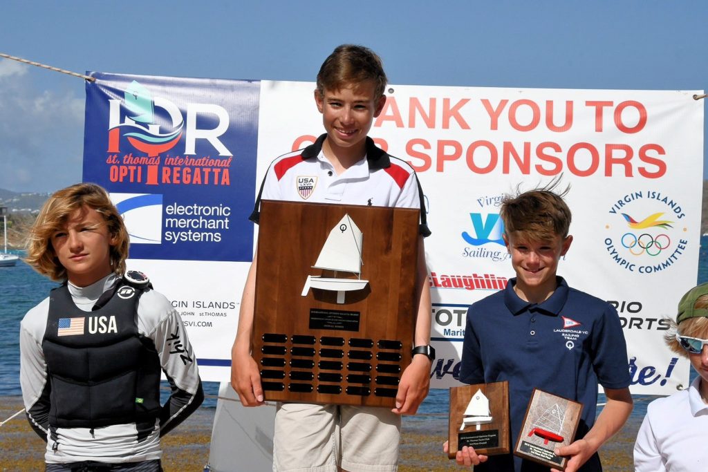 Three smiling young sailors stand in front of a 'Thank You to Our Sponsors' banner at a regatta. The center sailor holds a large trophy, while the others hold smaller awards. All are wearing sailing gear.