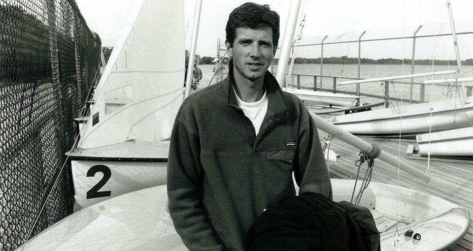A man wearing a jacket stands in front of sailboats docked by a chain-link fence, with water and land visible in the background. One boat is marked with the number 2. The image is in black and white.