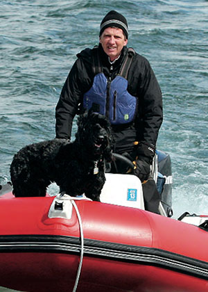 A man wearing a beanie and life jacket pilots a small red inflatable boat on the water, with a black dog standing at the front of the boat.