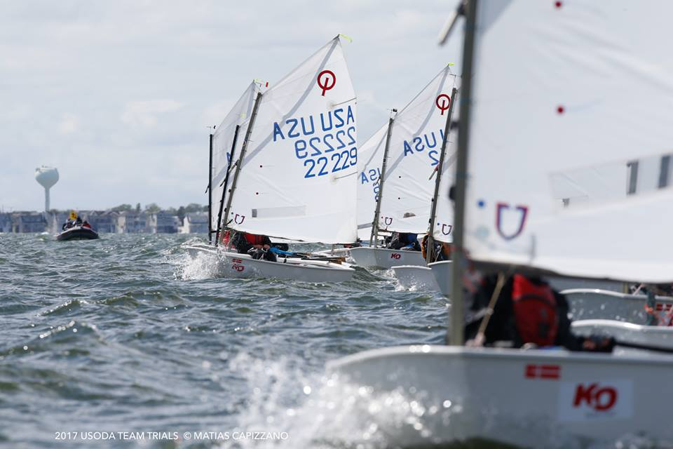 Several small sailboats with young sailors race on choppy water, their white sails displaying “USA” and boat numbers. A distant shoreline and a water tower are visible in the background under a cloudy sky.