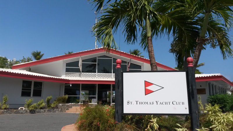 White building with red trim, surrounded by palm trees, features a sign in front reading 'St. Thomas Yacht Club' with a red, white, and black nautical flag logo under a clear blue sky.