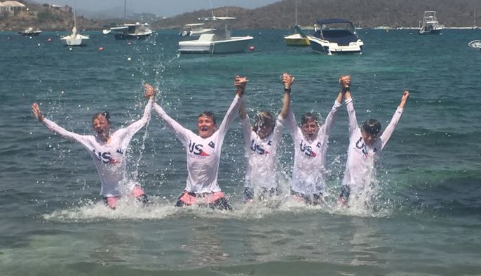 Five people wearing matching white 'USA' shirts stand in shallow ocean water, holding hands and raising their arms, splashing water joyfully. Boats and distant islands are visible in the background.