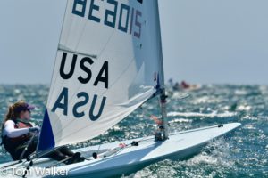 A sailor in a small sailboat marked "USA" and "BEA015" navigates choppy waters on a sunny day. The person wears sunglasses and a life vest. Other sailboats are visible in the background. © Tom Walker.