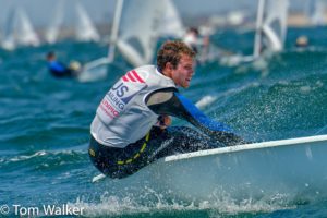 A male sailor in a "US Sailing" vest is intensely navigating a small sailboat on choppy blue water, with other sailboats and blurred sailors in the background. Photo credit reads "© Tom Walker.