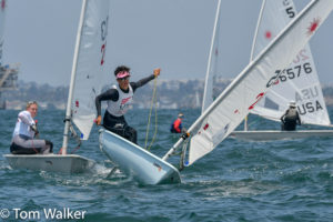 Several sailors compete in a dinghy race on choppy water, with one sailor in the foreground leaning out to balance the boat. Other boats and participants are visible in the background under a hazy sky.