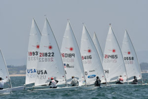 Several small sailboats with white sails, each marked with numbers and country codes, race closely together on the water under a clear sky. Sailors are focused on steering their boats.