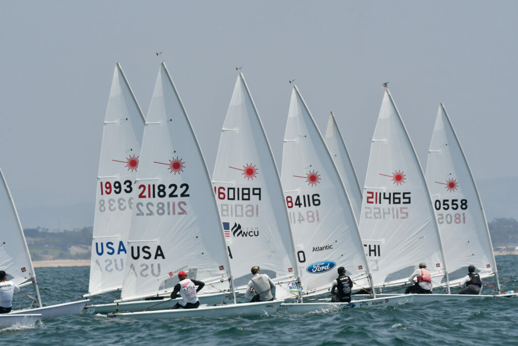 Several small sailboats with white sails, each marked with numbers and country codes, race closely together on the water under a clear sky. Sailors are focused on steering their boats.