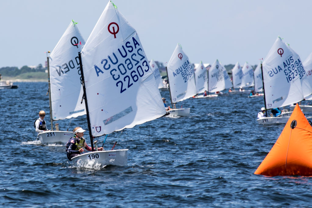 A group of young sailors in small sailboats race on open water toward an orange buoy, with visible sail numbers and a distant shoreline under a clear sky.