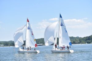 Two sailboats with white sails, marked with red and blue flags and numbers 6 and 3, race side by side on a calm lake with several people aboard. Trees and buildings are visible in the background under a clear sky.