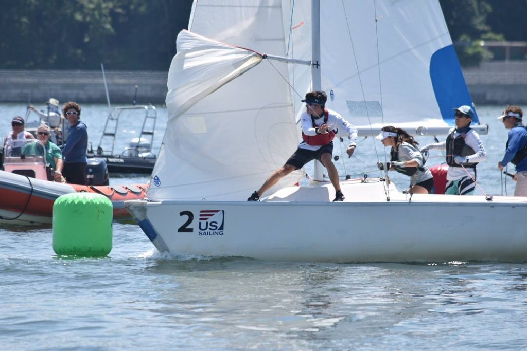 A sailboat with three crew members racing past a green buoy, marked '2 US Sailing,' as people watch from nearby boats on a sunny day.