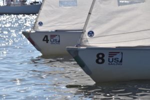 Two small sailboats with "US Sailing" logos and the numbers 4 and 8 on their bows are side by side on the water, with sunlight reflecting off the rippling surface.