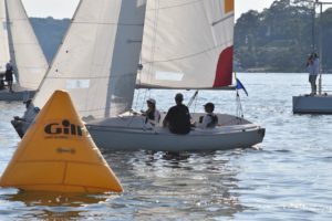 A small sailboat with three people passes a yellow buoy labeled "Gill" during a regatta on a sunny day, with other sailboats and a shoreline in the background.