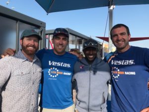 Four men stand close together smiling at the camera. Two wear matching blue "Vestas 11th Hour Racing" shirts, one wears a patterned button-up shirt and cap, and the fourth wears a gray jacket and sunglasses. They are outdoors under umbrellas.