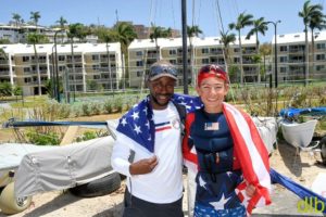 Two smiling sailors stand side by side, draped in American flags, in front of sailboats and palm trees with apartment buildings in the background on a sunny day.