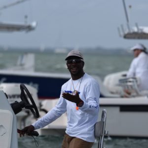 A man wearing sunglasses, a white shirt, and a cap smiles and gestures while steering a boat. There are other boats and a person in the background on a cloudy day.