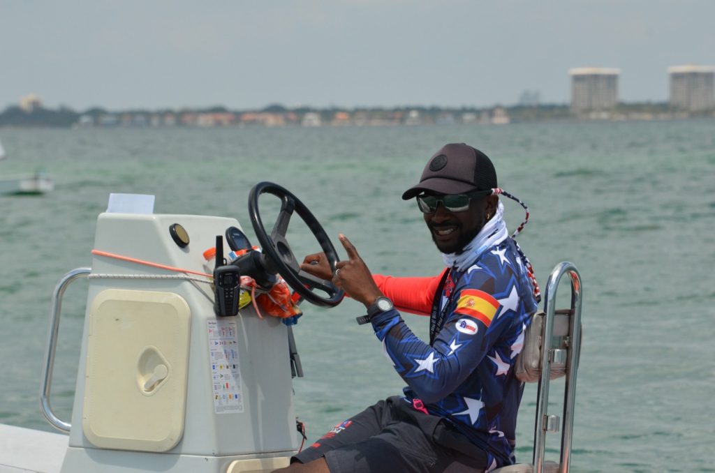 A man wearing sunglasses, a cap, and a star-patterned shirt smiles and gives a thumbs-up while steering a boat on the water, with a city skyline visible in the background.