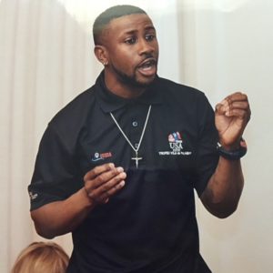 A man wearing a black polo shirt with USA logos and a cross necklace is speaking passionately, gesturing with his hands. He appears to be addressing an audience.