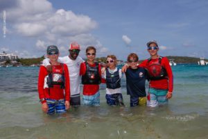Six smiling people, five kids and one adult, stand together in shallow, clear water at the beach, wearing life vests and sunglasses. The background shows blue sky, boats, and distant shoreline.