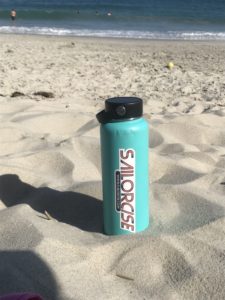 A turquoise water bottle with a "SAILORCISE" sticker rests on sandy beach with the ocean and swimmers in the background.