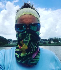 Person wearing sunglasses, a headband, and a colorful floral face covering, standing outdoors near water with trees, houses, and a cloudy sky in the background.