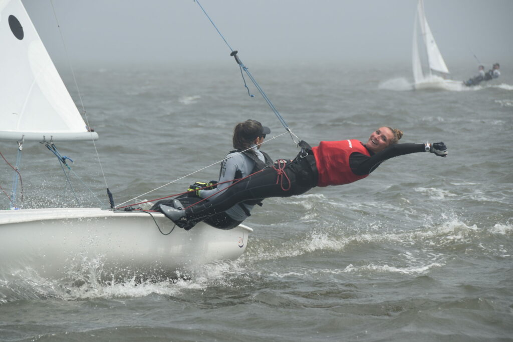 Two sailors in wetsuits and life jackets are on a small sailboat in choppy water; one leans far out over the edge, holding onto a rope, while another sailboat is visible in the background.