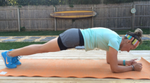 A person wearing a light blue shirt, gray shorts, and blue sneakers practices a plank on an orange mat outdoors. A wooden fence and a surfboard decoratively mounted are visible in the background.