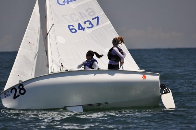 Two people wearing life vests sail a small white sailboat with the number 28 and sail number 6437 on the open water under a clear sky.