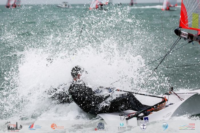 A sailor in a wetsuit is hit by a large splash of water while sailing a small boat in choppy waters; other sailboats and a cloudy sky are visible in the background. Various event logos appear at the bottom.