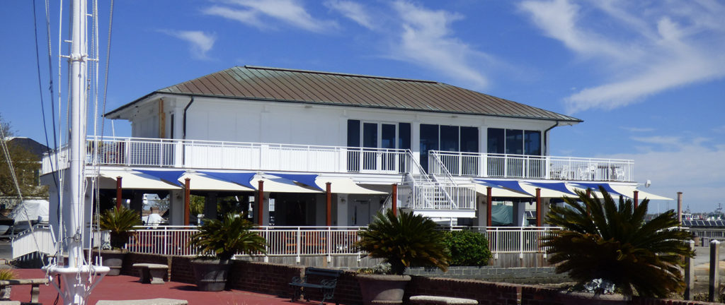 A two-story white building with large windows, a metal roof, and blue-and-white awnings, surrounded by palm trees and outdoor seating, under a blue sky with wispy clouds.