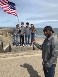 A man in a gray shirt and black cap points and smiles at four teens standing on rocks by the sea, one holding an American flag. The sky is partly cloudy and waves are visible in the background.